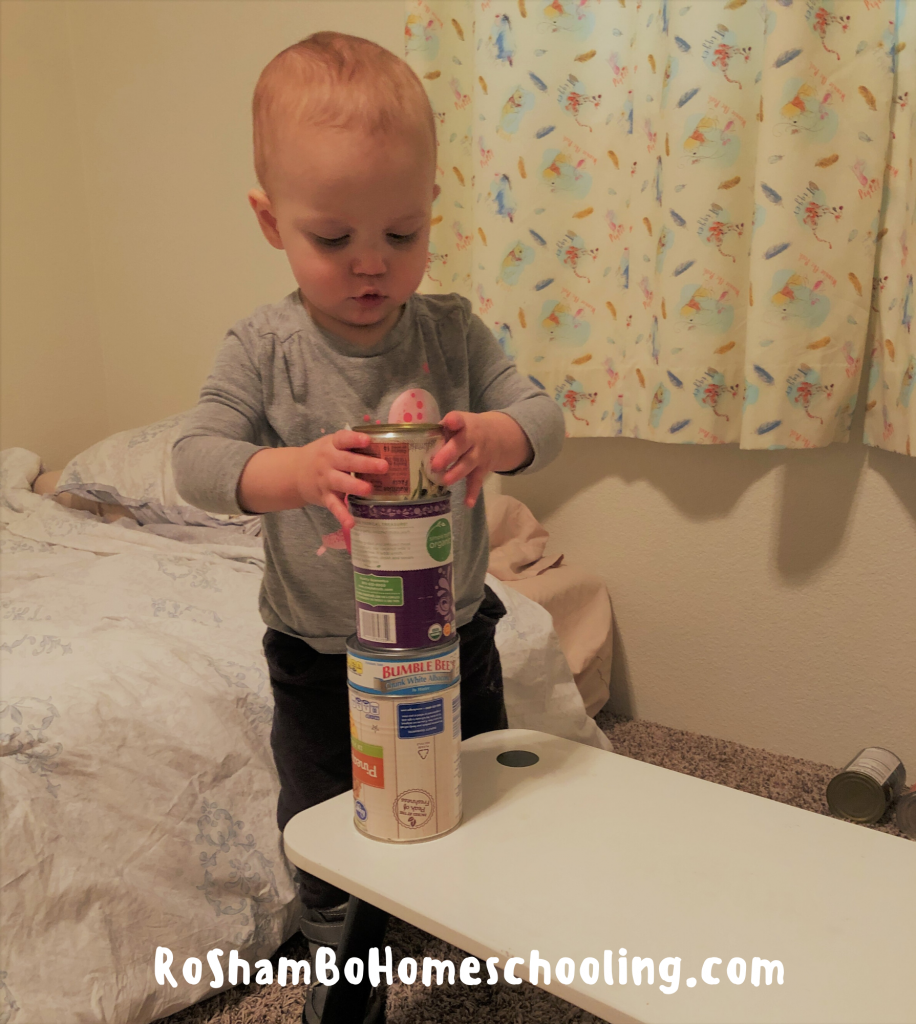 RoShamBo Homeschooling motor activity where a toddler is carefully stacking cans of food into a tower